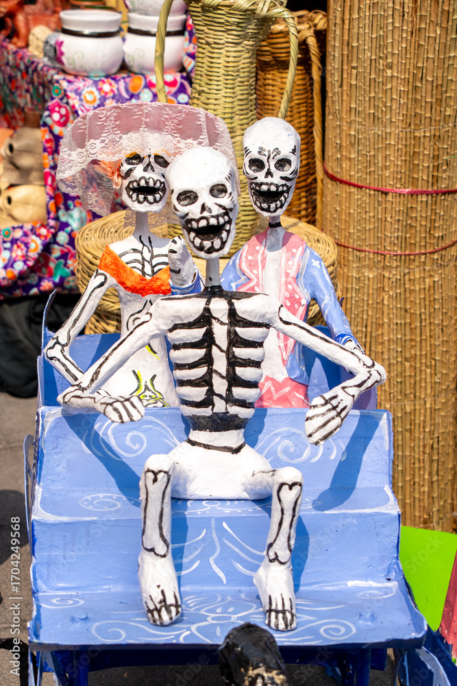 Fototapeta premium A skeleton is sitting on a blue bench with two other skeletons. The bench is decorated with skulls and bones. Day of the Dead celebration in Mexico, papel picado crafts, offerings, La Catrina
