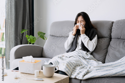 Young Asian woman get sick with changing seasons blowing nose while sitting on couch. Health problems, allergy, cold and flu concept. Copy space