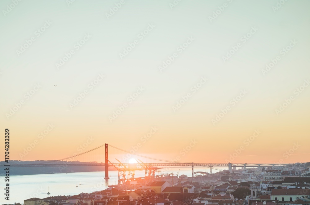 Obraz premium The sun sets behind the 25 de Abril Bridge in Lisbon, casting a warm golden glow over the Tagus river and the cityscape. Gentle light fills the sky and reflects on the water