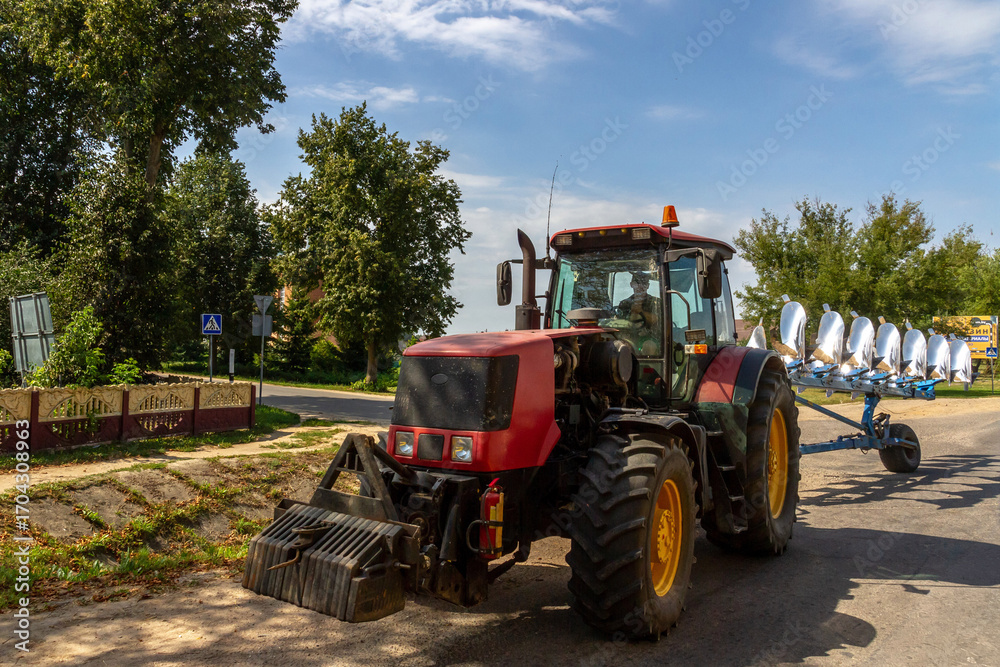 Obraz premium Agricultural tractor towing an 8-furrow plough in transport position on country road, showing curved moldboards for soil cultivation. Rural Belarus scene.
