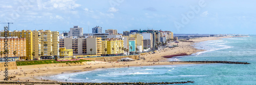 Wide panoramic view of the popular Playa de la Victoria in Cádiz, with its calm waters and golden sand in the sunlight, while the city buildings rise on the horizon