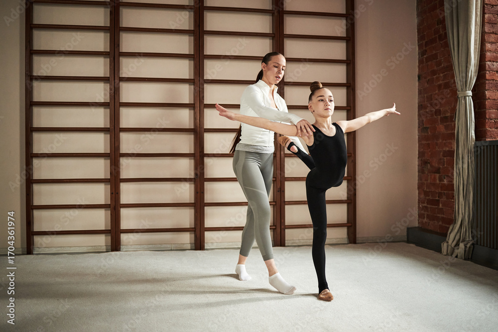 Fototapeta premium Caucasian young woman assisting child girl practicing ballet pose in dance studio, instructor supporting student during stretching exercise near wooden wall bars