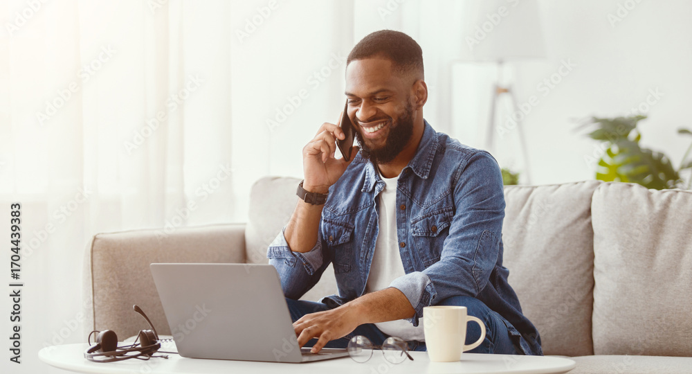 Naklejka premium Happy millennial african american male work on laptop and talk by phone at workplace on couch in living room interior. Success home business, distant communication and new normal due covid-19 pandemic