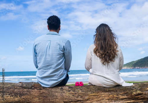 couple looking at the sea sitting on a tree trunk with baby shoes, first-time mother and father