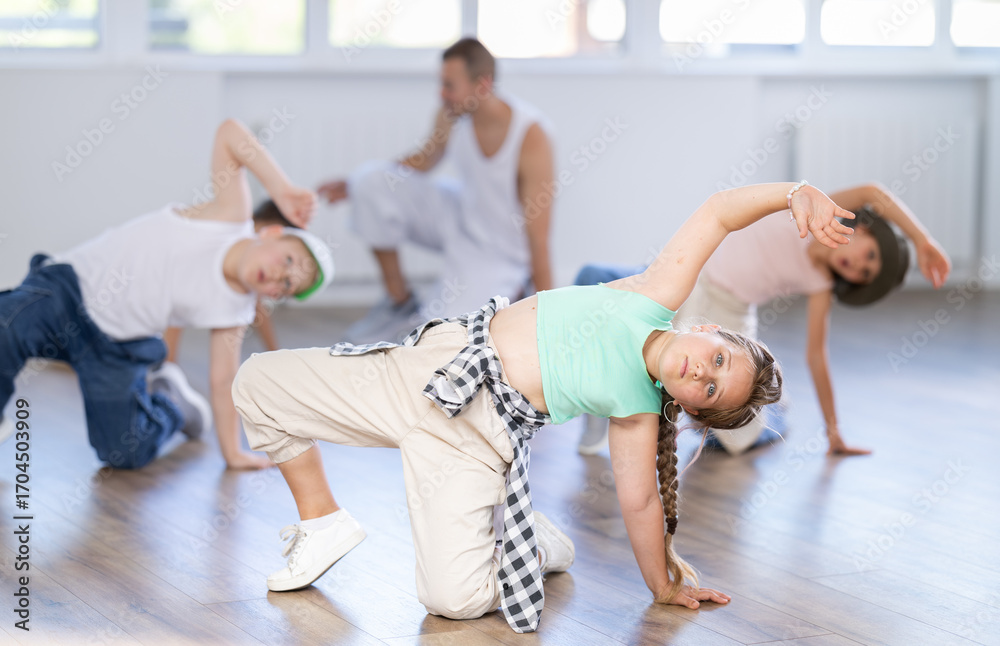 Fototapeta premium Preteen girl performing hip hop dance moves, showcasing skills and concentration, during group class for kids led by young male instructor in street dance studio..