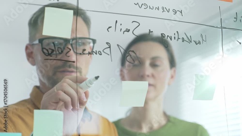 Business professionals brainstorming near glass board, using sticky notes for strategic planning, writing calculations, sharing innovative ideas during collaborative work session