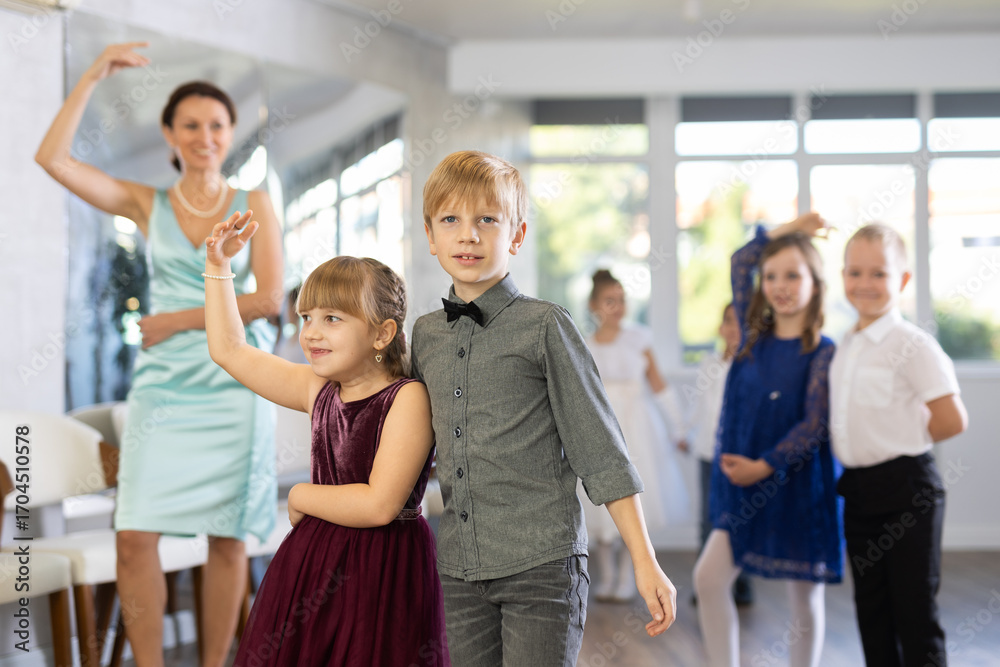 Obraz premium Smiling junior schoolchildren in party dresses learning dancing waltz with pedagogue in social hall
