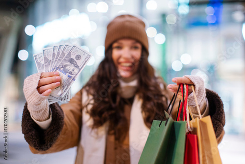 Happy woman holding shopper bags and fan of cash money banknotes, standing outdoors in front of shopping mall, purchasing for winter holidays