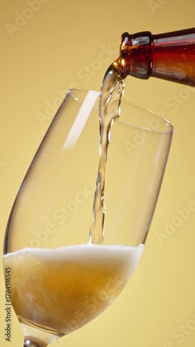 Cold craft light beer pouring in glass from bottle, slow motion close up. Stream wheat malt beverage flowing in clear goblet making dense bubbling foam. Froth heady drink foaming on yellow background