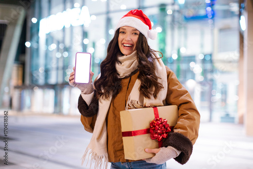 Xmas mobile offer. Excited lady showing blank cellphone screen and holding gift box, standing in front of shopping center, mockup for ad