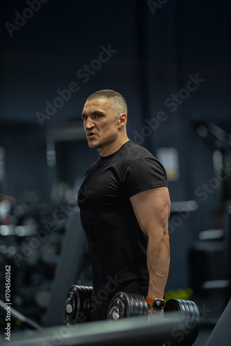 Strong athletic man stands focused in a gym gripping heavy dumbbells while preparing for an intense weightlifting session demonstrating fitness dedication.