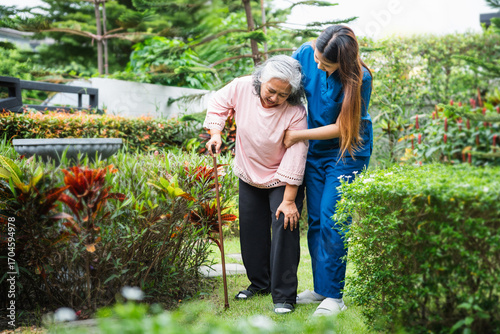 Senior care and assistance, Young caregiver helping senior woman to walk with a cane in the garden, Nurse helping an elderly patient for a walk outdoors