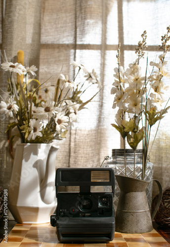 Bohemian image of an antique instant camera on an aged surface surrounded by vases of flowers with a window in the back