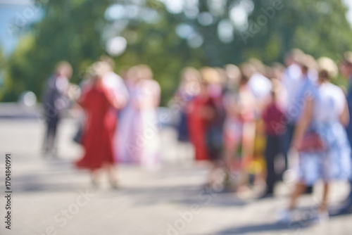 Blurred crowd of people socializing outdoors in sunlit urban setting with lush green trees.