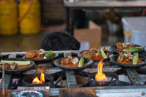 A close-up shot of several plates of Indonesian Ayam Penyet (smashed fried chicken with sambal) on a rustic street food stove. The flames below are heating the food for serving. Authentic outdoor cook