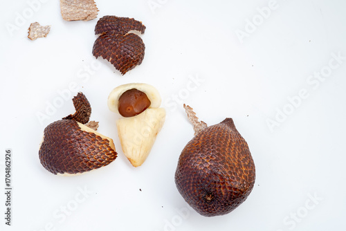 A few spiky brown salak fruit, one peeled to reveal the white flesh and a dark brown seed, are arranged on a clean white surface with some loose pieces of peel.