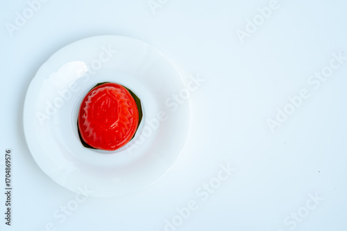 A top-down shot of a single red, oval-shaped Chinese sweet dumpling with a decorative pattern, sitting on a leaf on a white plate. The plate is positioned on a white background with ample negative spa
