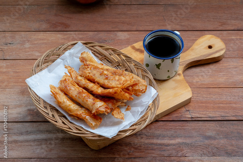 A woven plate holds several pieces of golden-brown fried banana and chocolate spring rolls, placed next to a mug of black coffee on a small wooden cutting board.