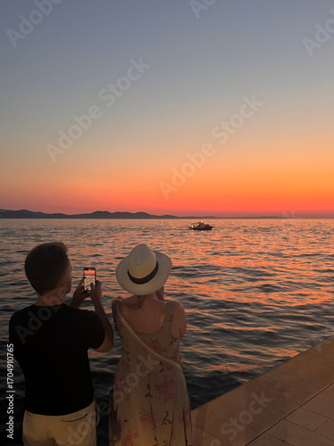 Fototapeta Naklejka Na Ścianę i Meble -  rear view of man and woman standing together taking photo watching sunset over Adriatic Sea in Zadar, Croatia