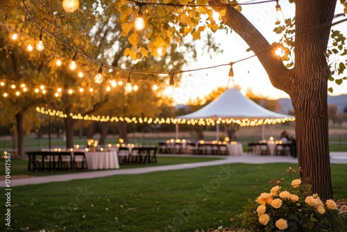 A beautifully decorated outdoor event space with string lights, a tent, and tables surrounded by vibrant autumn foliage.