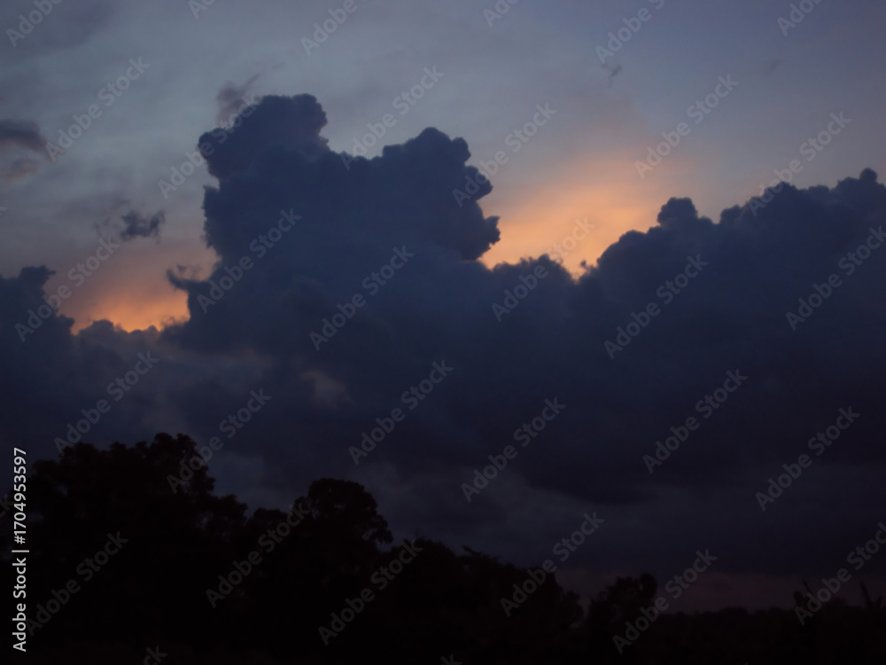 Fototapeta premium A dramatic, dark cloud formation silhouetted against the warm glow of a sunset sky above a treeline.