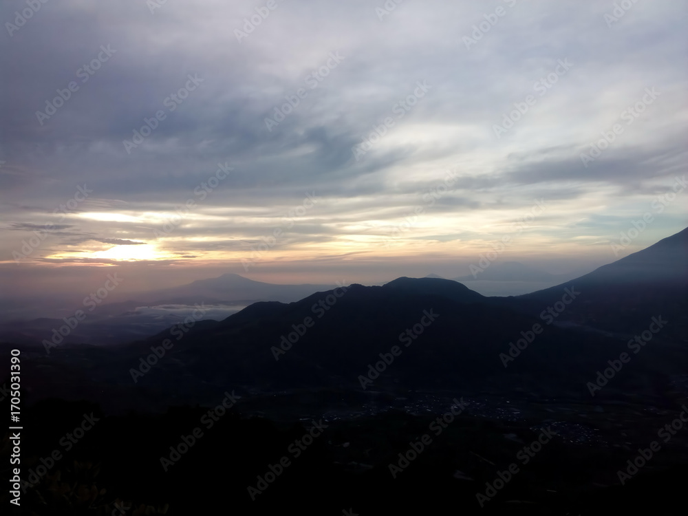 Obraz premium A dramatic sunrise over silhouetted mountain ranges under a cloudy, atmospheric sky.
