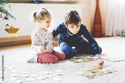 Two little chilren, cute toddler girl and school kid boy playing together card game by decorated Christmas tree. Happy healthy siblings, brother and sister having fun together. Family celebrating xmas