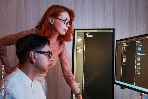 Two colleagues analyze code on dual monitors in a modern office, collaborating on software development, debugging, and data review at a workstation.