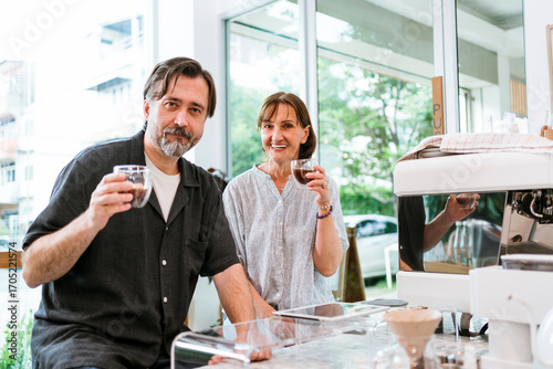 Smiling adults enjoying espresso in a bright modern cafe beside a professional coffee machine, raising cups at the counter with natural light and urban view through large windows.