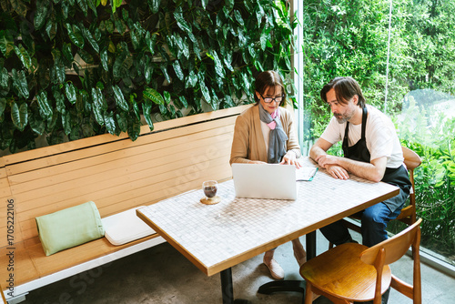 Two colleagues in a cafe with a laptop discuss work beside a lush indoor green wall, suggesting consulting or small business planning in a modern, casual workspace.