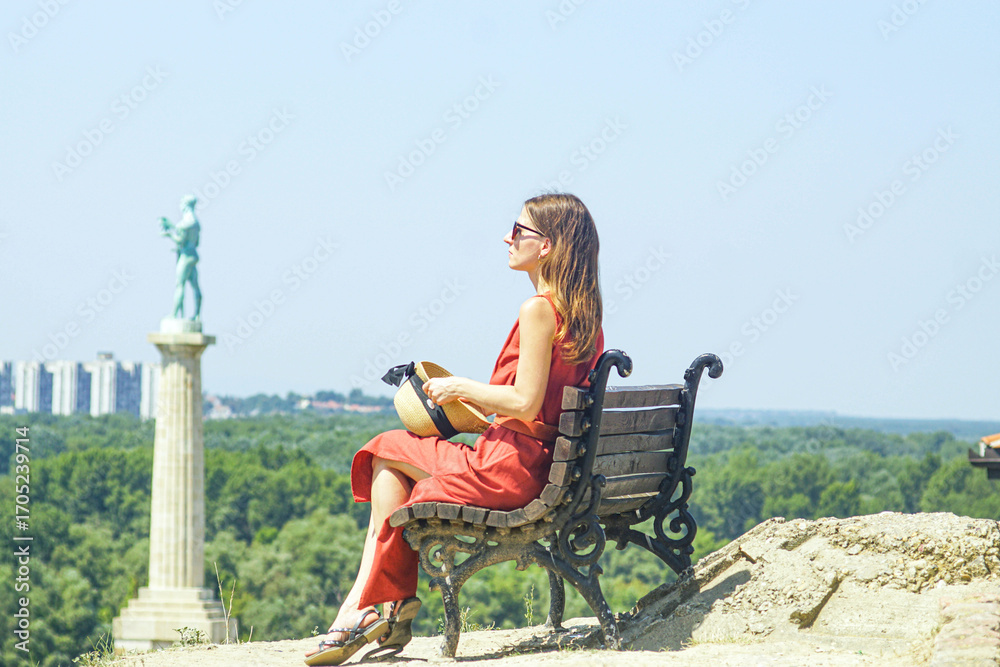 Fototapeta premium Stylish woman admiring panoramic view from Belgrade Fortress with iconic Pobednik monument background. Cultural tourism in Eastern Europe. Authentic travel experience and heritage site exploration