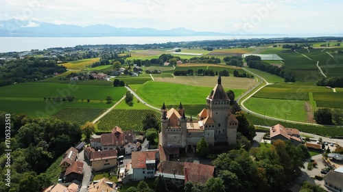 Aerial view of Château de Vufflens, a medieval castle rising amidst lush green fields and vineyards, contrasting with the distant blue lake, Vufflens-le-Château, Vaud, Switzerland.