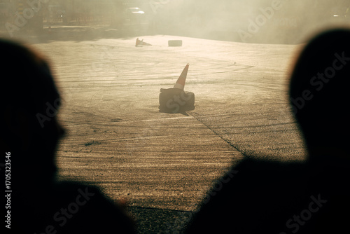 Golden light, smoke, and silhouettes of spectators create a dramatic scene at a drift show, with cones and skid marks on the asphalt.
