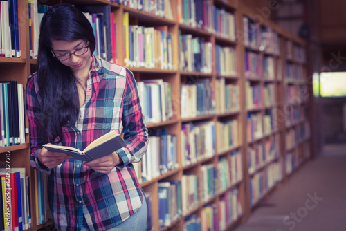 Student leaning against tall wooden bookshelf in library aisle, reading open book with glasses