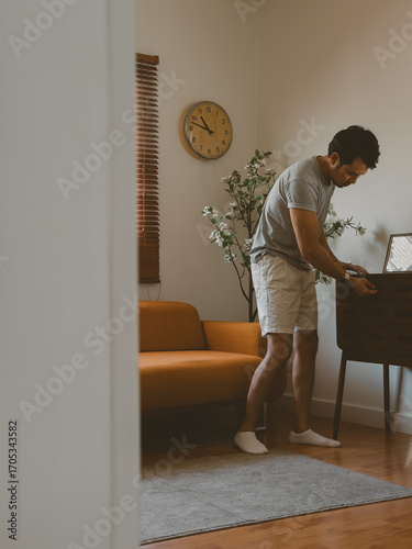 Young Thai man bending over dining table arranging items in warm indoor light in a quiet minimalist Thai apartment with natural daylight