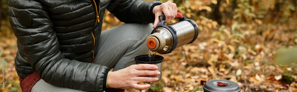 Naklejka premium Senior woman enjoys a warm drink while hiking in a vibrant forest setting on a cool day