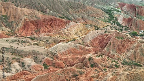 Aerial footage of Skazka Canyon in Kyrgyzstan as the drone steadily ascends, revealing the vast red rock formations and stunning mountain backdrop in warm daylight.