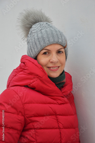 Portrait of pretty Caucasian woman in red winter jacket and grey knitted hat with fur pompom on white background. Women's warm street clothes.
