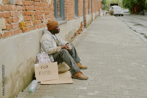 Bild auf Leinwand Black middle aged man sitting on sidewalk leaning against brick wall holding cup
