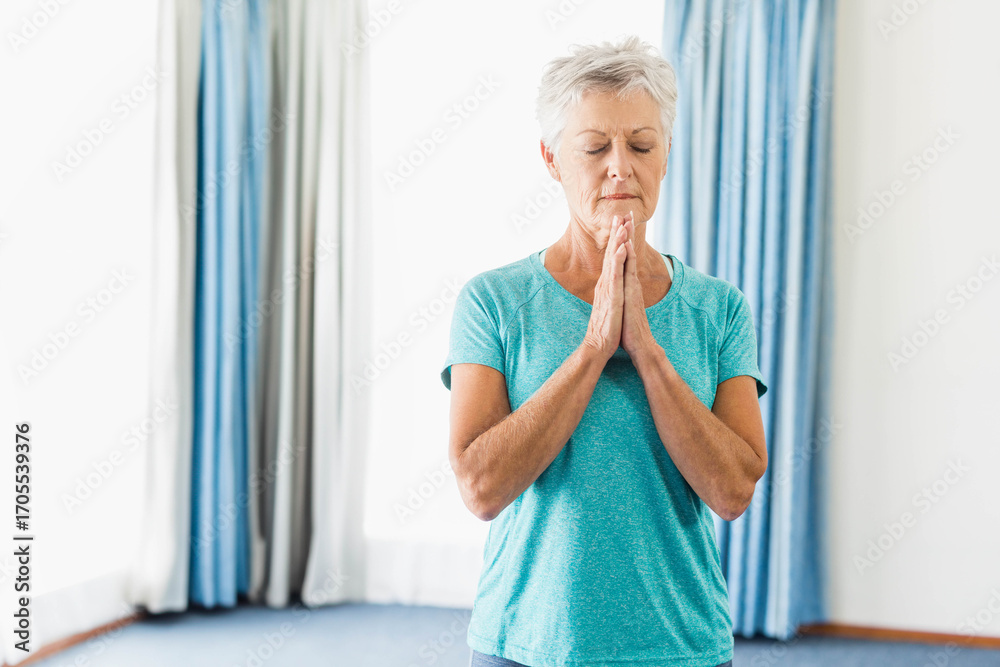 Naklejka premium Senior woman standing next to tall curtains in home studio meditating in teal workout shirt