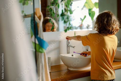 A child stands at a stylish bathroom sink, brushing their teeth. Morning light fills the space, creating a fresh and inviting atmosphere with greenery in view.