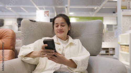 Young woman seated comfortably on armchair using smartphone, immersed in digital communication and social media, relaxed modern lifestyle in spacious indoor setting with technology, connection 