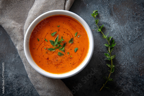 Creamy tomato soup garnished with fresh herbs in white bowl on dark rustic background, homemade red vegetable cream soup with parsley topping, healthy warm comfort food meal for lunch dinner