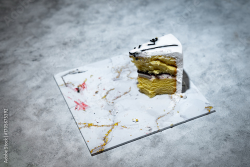A tart cake slice with layers of yellow sponge cake, white cream, and fruit jam in the middle is placed on a marble patterned base.