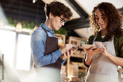 Low angle view of two concentrated female assistants in aprons working in small organic store. They standing together and fixing prices of new products, using tablet computer and notepad.