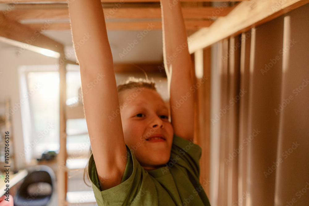 Fototapeta premium A young boy plays indoors, demonstrating strength while hanging from a wooden structure. Nearby, interactions with his father and brother occur, showcasing family bonding moments during the day.