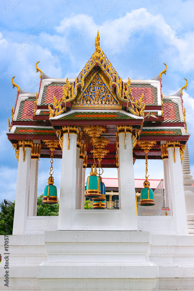 Naklejka premium Wat Rakhang temple pavilion in Bangkok, Thailand. White columns and golden Thai motifs frame hanging pastel blue green bells under bright blue sky, iconic Buddhist landmark.