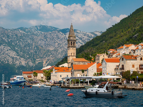 Sailing boats and waterside stone buildings on the Bay of Kotor shoreline at Perast in Montenegro. Rugged mountains in the background. Clear blue sky.