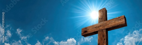 A wooden cross stands against a bright blue sky dotted with fluffy white clouds, with the sun's rays shining brightly from behind it