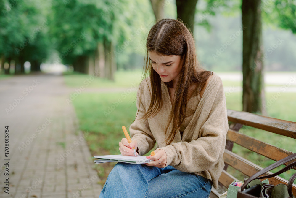 Obraz premium A young woman sits on a wooden bench in a scenic park, focused on writing in her notebook. Lush green trees surround her, creating a serene atmosphere.